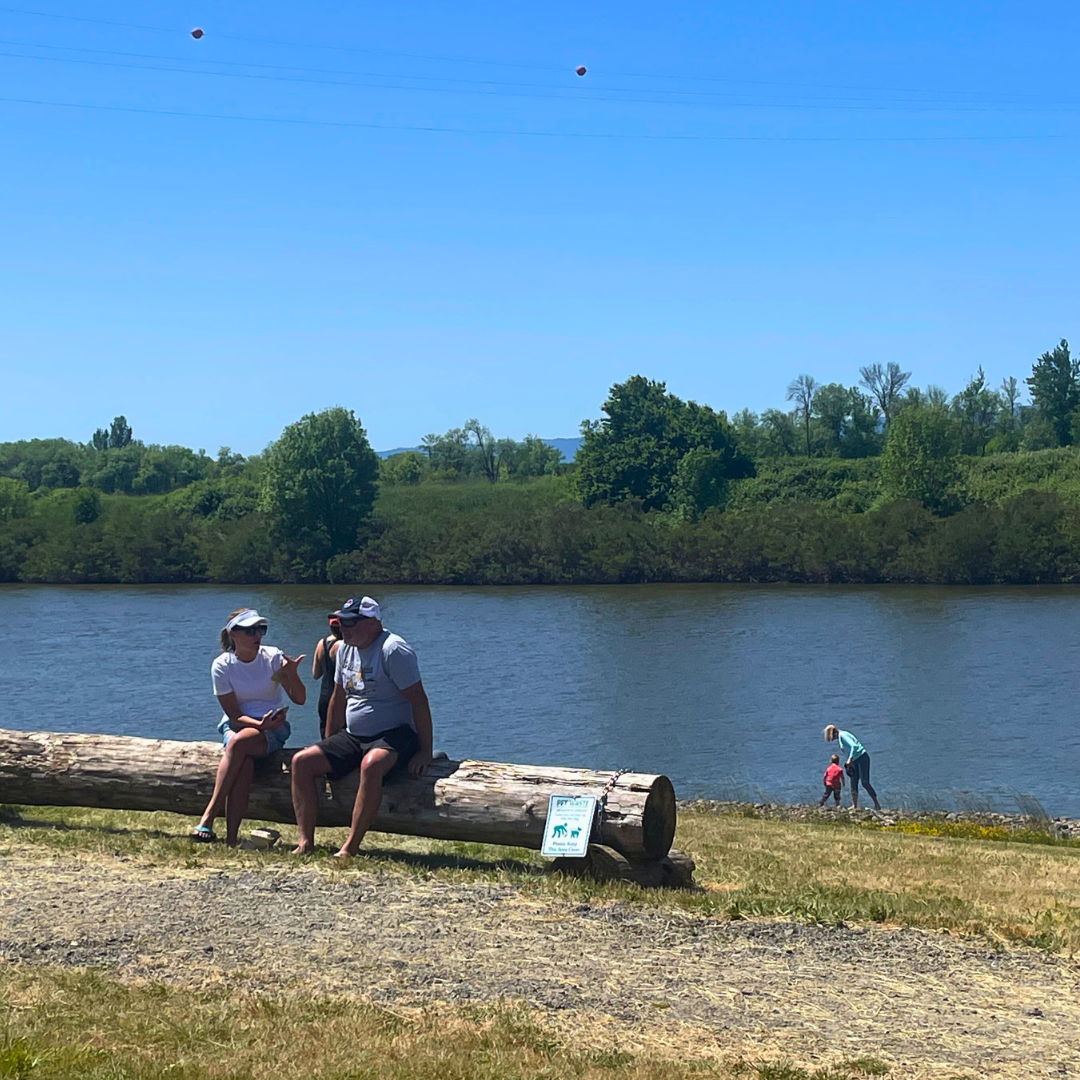 People conversing on a log bench at the Ridgefield Waterfront with a child and parent wading into La