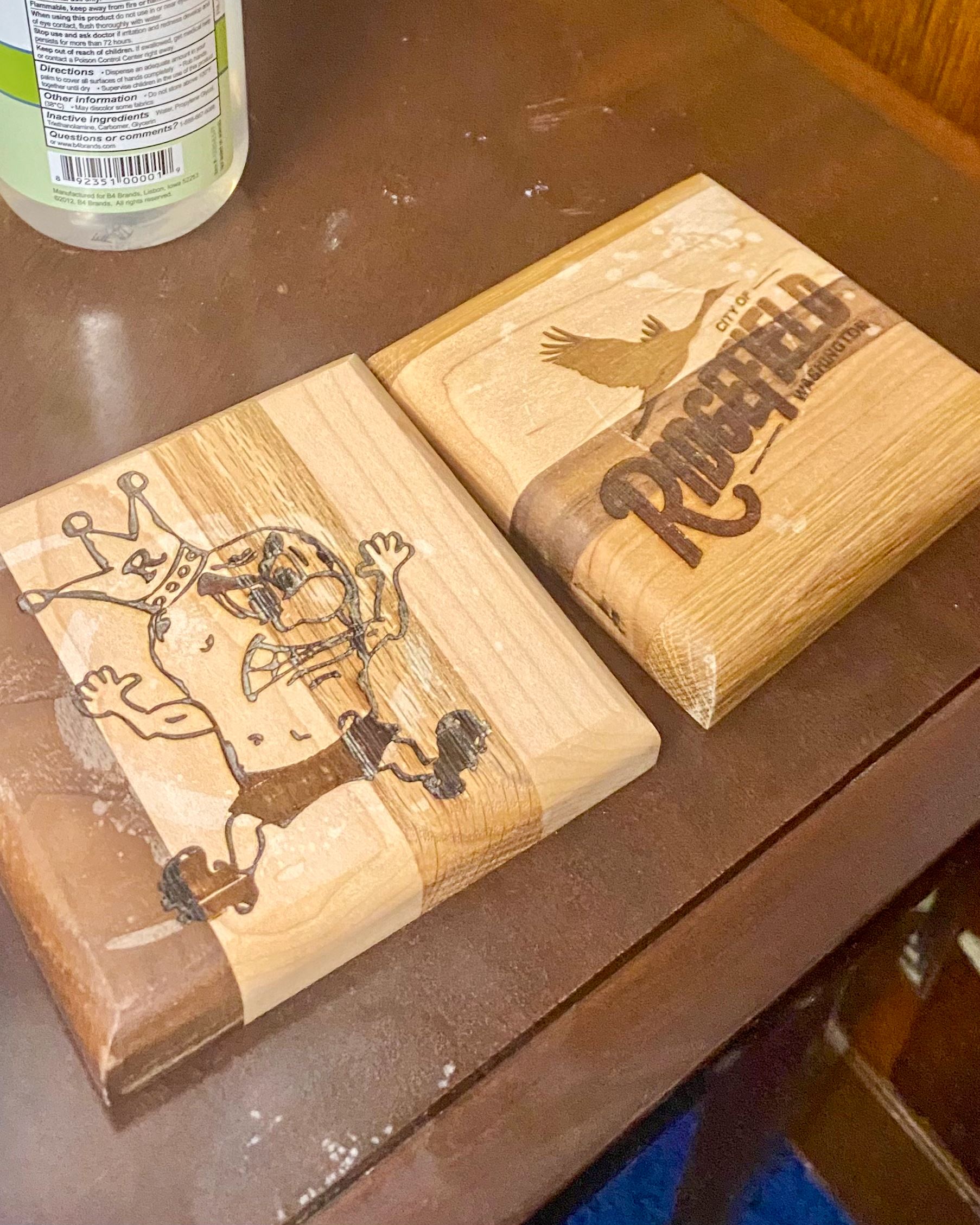 Two wooden coasters with burned designs of the Ridgefield city logo and the Spudder mascot on them.